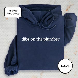 A navy blue hoodie with the text "dibs on the plumber" printed on it, along with a speech bubble indicating that the hoodie is available.