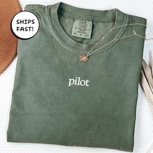 May include: An olive green t-shirt with the word "pilot" embroidered in white. A gold-coloured necklace with a pendant rests on the shirt. The shirt is folded and has a tag that says "Comfort Colors".