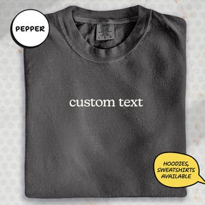 A gray t-shirt with the text "custom text" printed on it, along with a label indicating it is a product from the "Pepper" brand. The t-shirt is folded and displayed on a white background, with a speech bubble indicating that hoodies, sweatshirts, and other items are available.