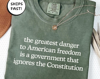 Puede incluir: Camiseta verde oliva con el texto "el mayor peligro para la libertad estadounidense es un gobierno que ignora la Constitución". Un collar dorado con un colgante descansa sobre la camiseta.