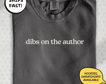 May include: Dark gray t-shirt with the phrase "dibs on the author" in white text. The shirt has a crew neck and a tag at the back of the neck. A speech bubble graphic says "Hoodies, Sweatshirts Available".