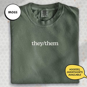 May include: A moss green t-shirt with the white text "they/them". The shirt is folded and has a Comfort Colors label. A speech bubble indicates hoodies and sweatshirts are available.