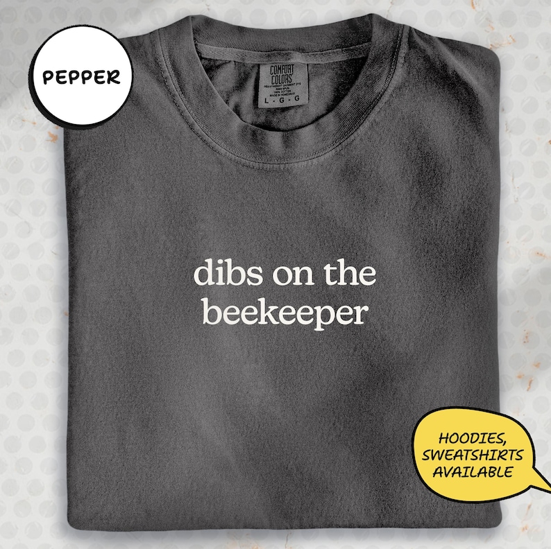 A gray t-shirt with the text "dibs on the beekeeper" printed on it, along with a label indicating it is from the "CITY COLORS" brand. The t-shirt is folded and displayed on a surface with a speech bubble indicating that hoodies and sweatshirts are available.