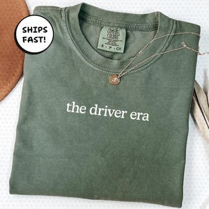 May include: An olive green t-shirt with the text "the driver era" in white. A gold-coloured necklace with a small pendant is draped across the shirt. The shirt is folded and has a tag that says "Comfort Colors".