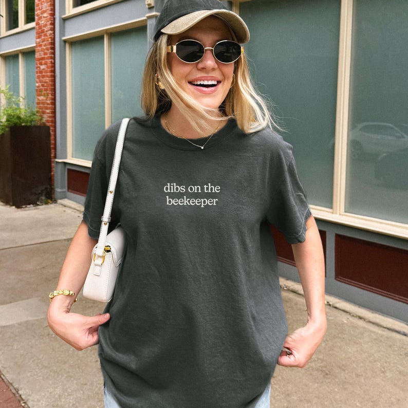 A smiling woman wearing a gray t-shirt with the text "dibs on the beekeeper" and sunglasses stands in front of a building.