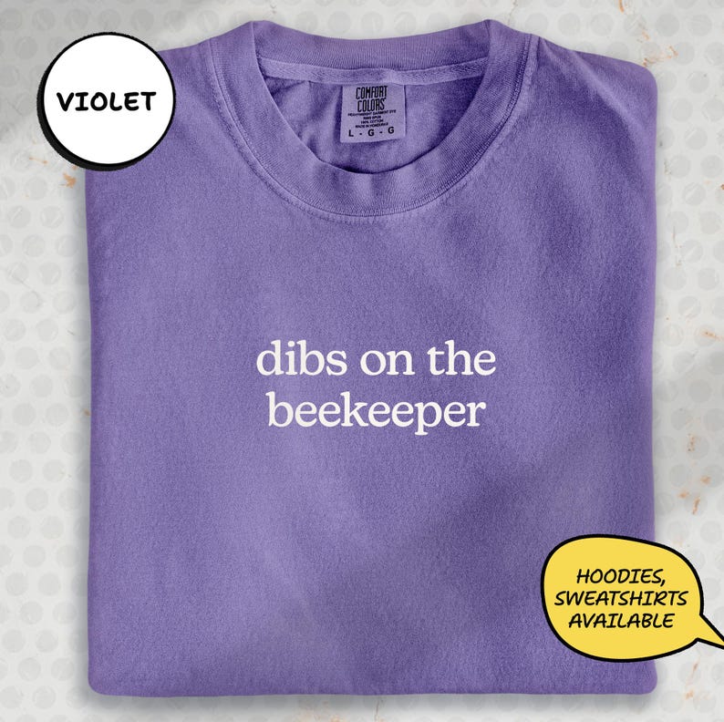 A purple t-shirt with the text "dibs on the beekeeper" printed on it.
