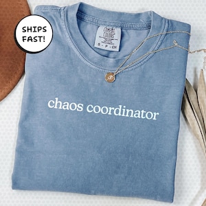 以下が含まれることがあります： 「chaos coordinator」の白い文字が入った青いComfort ColorsのTシャツ。ペンダント付きのゴールドネックレスがシャツの上にあります。シャツは白い表面にきれいに折りたたまれています。