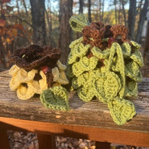 May include: Two handmade crochet items resembling autumn foliage. One is a brown and yellow flower, the other is a green and brown cluster of leaves. Both have a wooden bead center and are set against a blurred outdoor background.