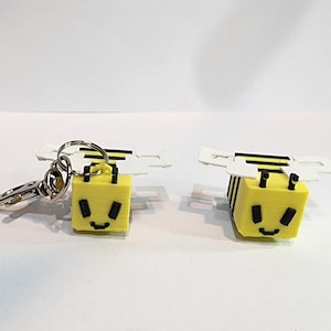May include: Two yellow cube-shaped bee keychains with black stripes and smiling faces. Each bee has white wings and a silver keychain clasp. One keychain is attached to a silver clasp.