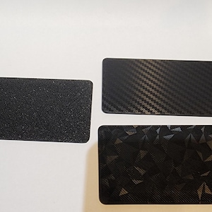 May include: Three black rectangular objects with textured surfaces. One has a speckled appearance, another a carbon fiber weave, and the third a geometric pattern. The objects are displayed on a white surface.
