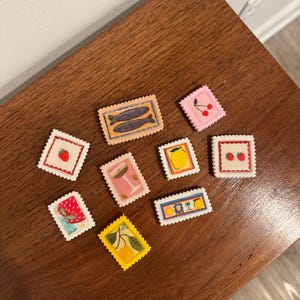 May include: A collection of colorful, decorative stamps with various designs, including fish, cherries, strawberries, lemons, and cocktails. The stamps are arranged on a dark wooden surface.
