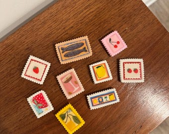 Op de afbeelding: Een verzameling kleurrijke, decoratieve postzegels met diverse ontwerpen, waaronder vissen, kersen, aardbeien, citroenen en cocktails. De postzegels zijn gerangschikt op een donker houten oppervlak.