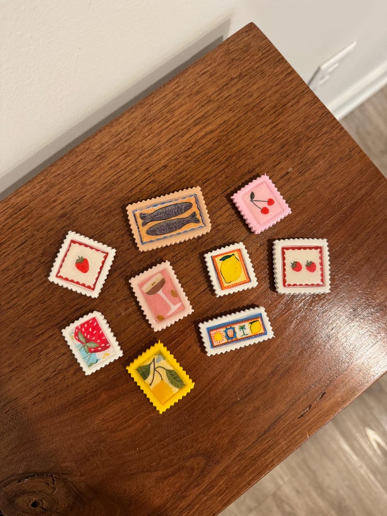 May include: A collection of colorful, decorative stamps with various designs, including fish, cherries, strawberries, lemons, and cocktails. The stamps are arranged on a dark wooden surface.