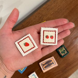 May include: Several decorative stamps with various designs are displayed. Two stamps feature strawberries, one with a single berry and the other with two. Other stamps show fish, a floral design, and a green square with a design.