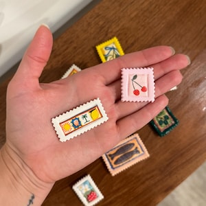 May include: A collection of miniature, decorative stamps with various designs, including cherries, fish, and tropical scenes. The stamps are rectangular with a scalloped edge and are in various colors, such as pink, yellow, and blue. The stamps are held in a hand.