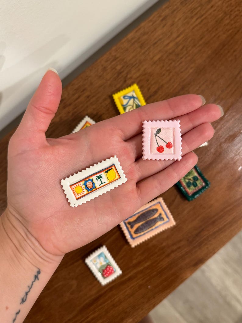 May include: A collection of miniature, decorative stamps with various designs, including cherries, fish, and tropical scenes. The stamps are rectangular with a scalloped edge and are in various colors, such as pink, yellow, and blue. The stamps are held in a hand.