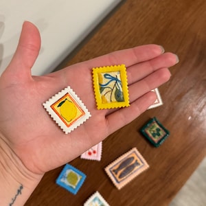 May include: A collection of miniature, colorful, postage stamp-shaped art pieces. The stamps feature various designs, including a lemon, a sprig of leaves, and a pair of fish. The stamps are in a variety of colors, including yellow, white, blue, and pink.
