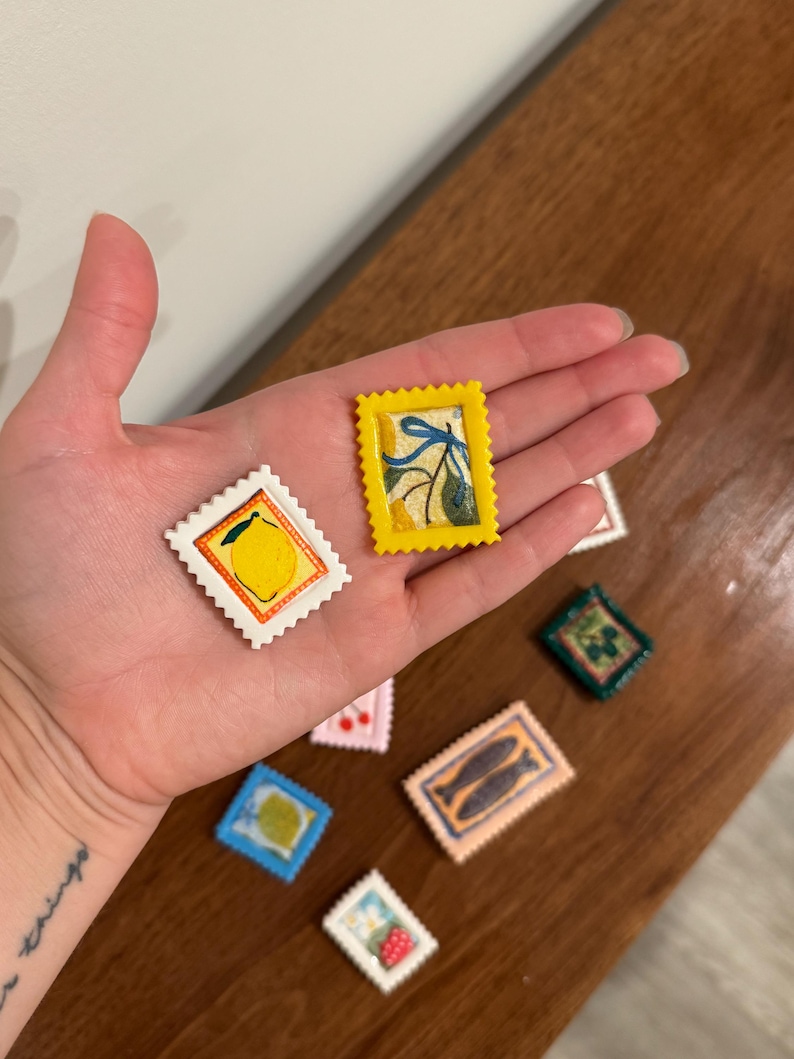 May include: A collection of miniature, colorful, postage stamp-shaped art pieces. The stamps feature various designs, including a lemon, a sprig of leaves, and a pair of fish. The stamps are in a variety of colors, including yellow, white, blue, and pink.