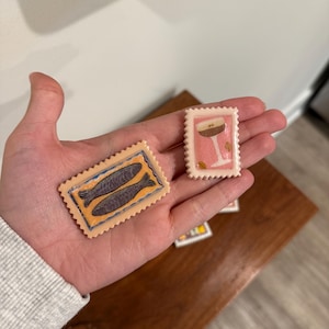 May include: Two decorative postage stamps are held in a hand. One stamp features two fish in a blue and orange design. The other stamp has a pink background with a cocktail glass design. The stamps have a serrated edge.