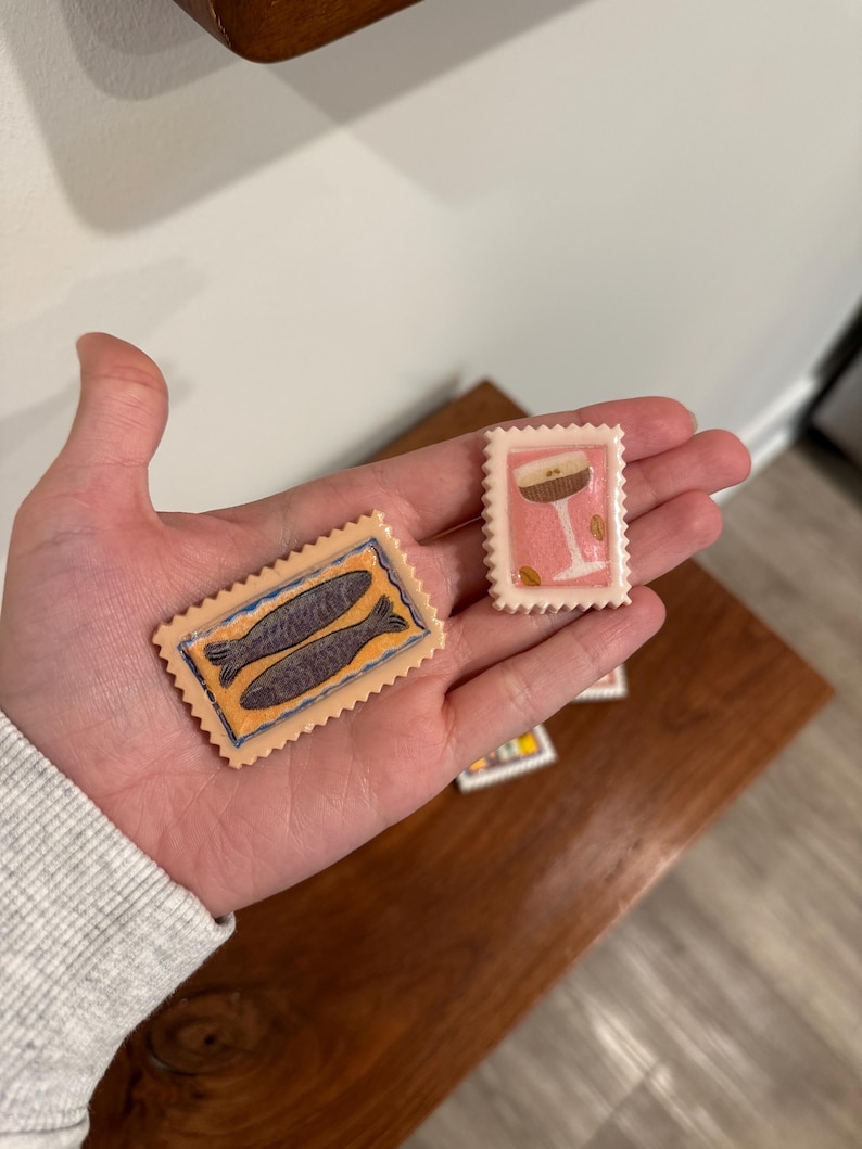 May include: Two decorative postage stamps are held in a hand. One stamp features two fish in a blue and orange design. The other stamp has a pink background with a cocktail glass design. The stamps have a serrated edge.