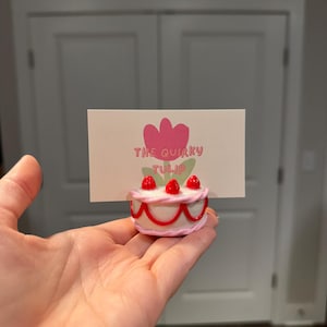 May include: A small, white cake-shaped card holder with pink and red accents, topped with three red berries. The card features a pink tulip graphic and the text "THE QUIRKY TULIP".