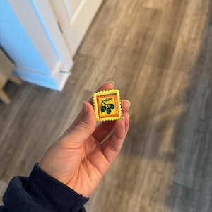 May include: A small, yellow stamp with a green olive branch design. The stamp has a red border and is held in a hand. The background is a wooden floor.