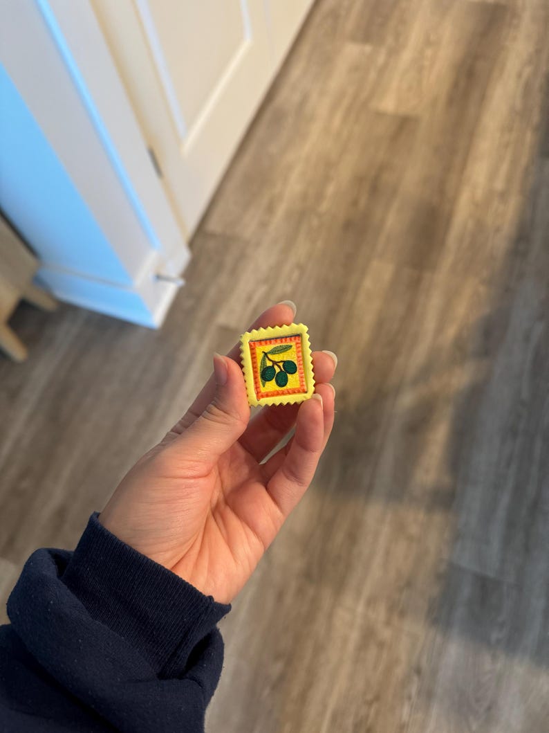 May include: A small, yellow stamp with a green olive branch design. The stamp has a red border and is held in a hand. The background is a wooden floor.