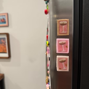 May include: Three decorative refrigerator magnets in the shape of postage stamps. Each stamp features a cocktail glass design with a brown drink and a white stem. The stamps are in shades of pink and tan.