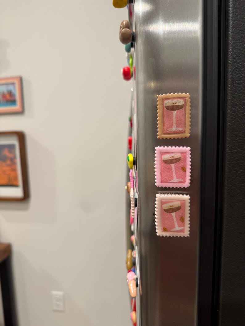 May include: Three decorative refrigerator magnets in the shape of postage stamps. Each stamp features a cocktail glass design with a brown drink and a white stem. The stamps are in shades of pink and tan.
