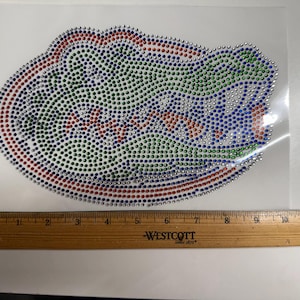 May include: A rhinestone transfer featuring an alligator head design in green, blue, orange, and clear crystals. A wooden Westcott ruler is positioned below the design, showing the transfer is approximately 11.5 inches wide.