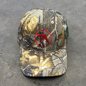 May include: Camouflage baseball cap with a red logo. The hat features a classic six-panel design with a curved brim. The camouflage pattern includes shades of brown, green, and tan, creating a naturalistic appearance. The logo is a stylized red design within a red circle.