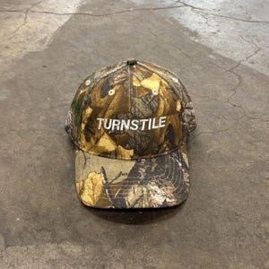 May include: A camouflage baseball cap with the word "TURNSTILE" embroidered in white across the front. The cap features a curved brim and a classic six-panel design. The hat is made of a durable fabric and is suitable for outdoor activities.
