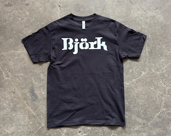 May include: Black short-sleeve t-shirt with the name "Björk" printed in large, white, stylized letters across the chest. The shirt is made of a soft material and has a crew neck.