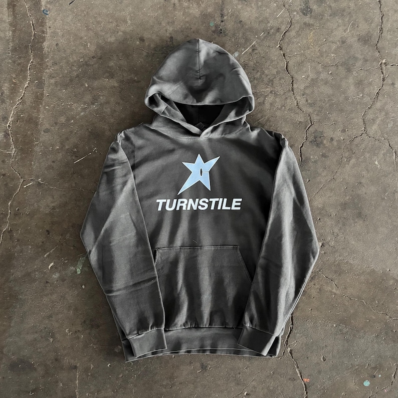 May include: A gray hoodie with a front pocket and a hood. The front of the hoodie features a light blue star graphic above the word "TURNSTILE" in white, sans-serif font. The hoodie is made of a soft material.