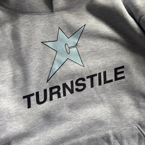 May include: A gray hoodie with a light blue star graphic and the word "TURNSTILE" in black block letters. The hoodie has a front pocket and a hood. The star graphic has a unique design.
