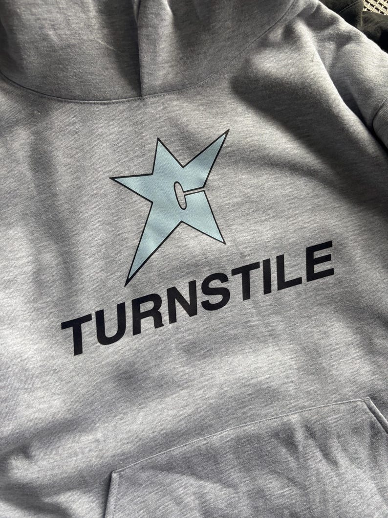 May include: A gray hoodie with a light blue star graphic and the word "TURNSTILE" in black block letters. The hoodie has a front pocket and a hood. The star graphic has a unique design.