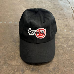 Könnte beinhalten: Schwarze Baseballkappe mit gebogenem Schirm. Die Kappe hat ein gesticktes Logo in Weiß und Rot. Das Logo ist eine stilisierte Wortmarke. Die Kappe besteht aus einem strapazierfähigen Stoff und hat einen Knopf oben.