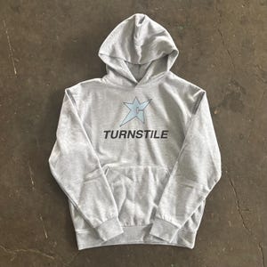May include: A light gray hoodie with a front pocket and a hood. The hoodie features a light blue star graphic and the word "TURNSTILE" in black text. The hoodie is made of a soft, comfortable material.