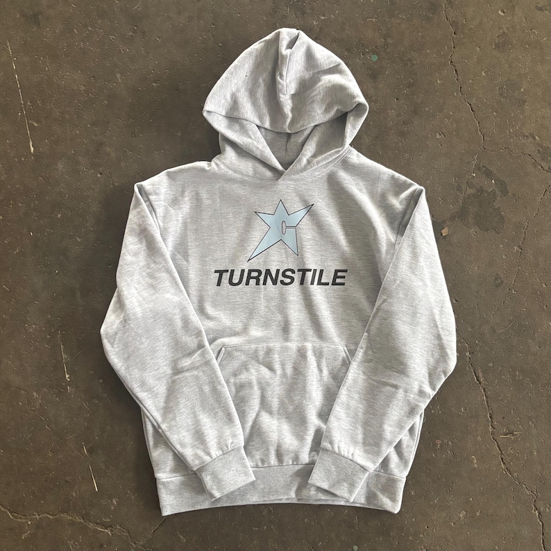 May include: A light gray hoodie with a front pocket and a hood. The hoodie features a light blue star graphic and the word "TURNSTILE" in black text. The hoodie is made of a soft, comfortable material.