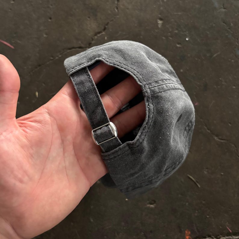 May include: A dark gray, slightly distressed baseball cap with a curved brim. The cap features a fabric strap with a silver-colored metal buckle for adjustment. The cap is made of a soft, worn material.