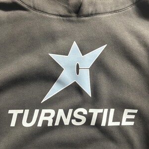 May include: A gray hoodie with a light blue star-shaped graphic and the word "TURNSTILE" in white block letters. The hoodie has a drawstring hood and a soft, textured fabric.