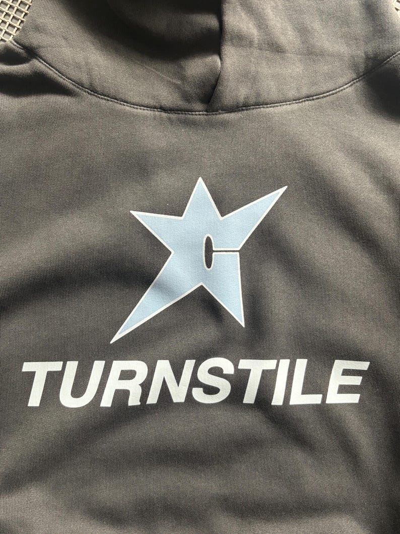 May include: A gray hoodie with a light blue star-shaped graphic and the word "TURNSTILE" in white block letters. The hoodie has a drawstring hood and a soft, textured fabric.
