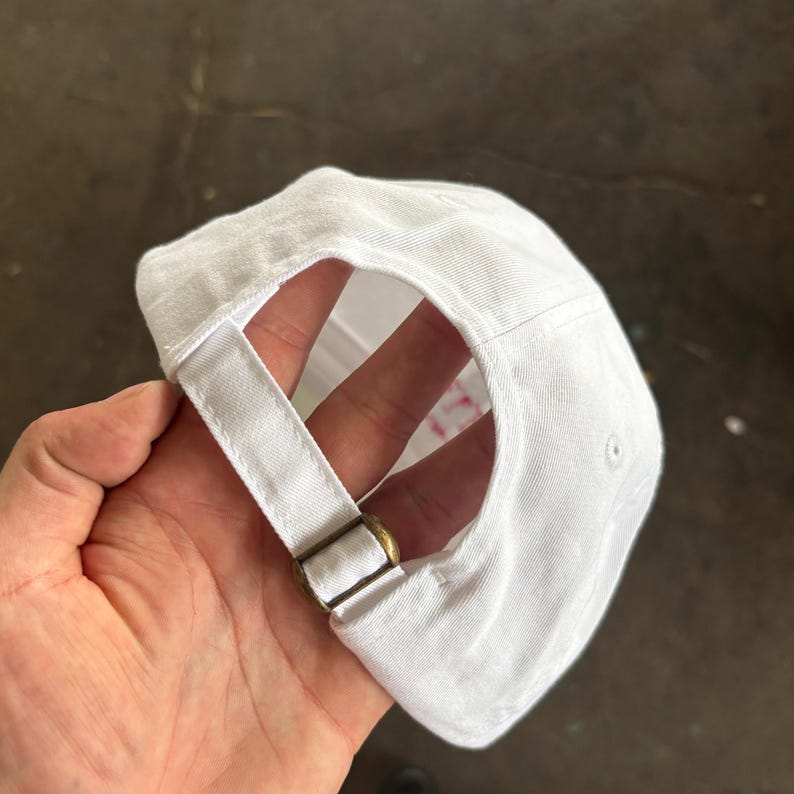 Puede incluir: Gorra de b&eacute;isbol blanca con visera curva y correa ajustable. La gorra tiene una hebilla met&aacute;lica de color lat&oacute;n en la correa para ajustar la talla. El interior de la gorra es visible, mostrando el forro y las costuras.