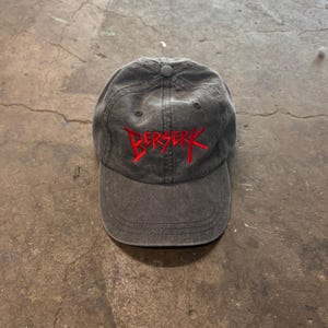 May include: A charcoal gray baseball cap with the word "BERSERK" embroidered in red across the front. The cap has a curved brim and a button on top. The hat is made of a soft, worn material.