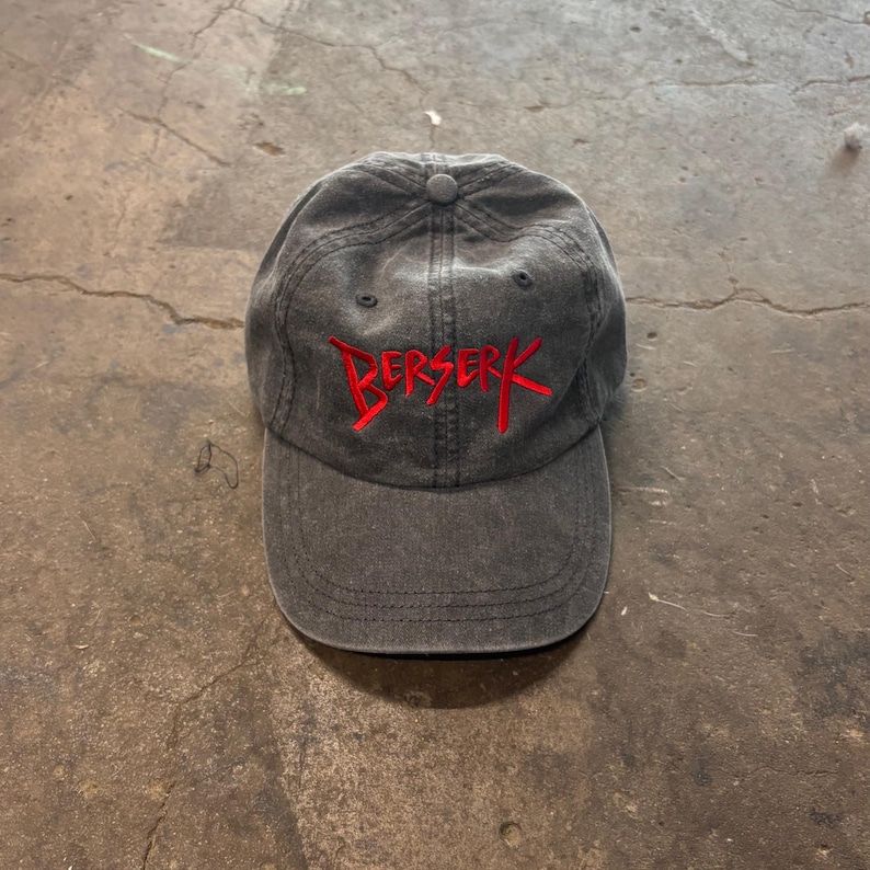 May include: A charcoal gray baseball cap with the word "BERSERK" embroidered in red across the front. The cap has a curved brim and a button on top. The hat is made of a soft, worn material.