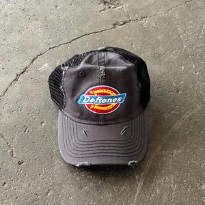 May include: Gray baseball cap with a black mesh back and distressed details. The cap features the band name "Deftones" in a blue, red, and yellow logo on the front. The hat is made of a durable fabric and has a curved brim.