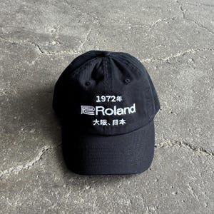 Puede incluir: Una gorra de béisbol negra con texto bordado en blanco que dice 1972年, Roland y 大阪、日本. La gorra tiene una corona estructurada, una visera curva y una costura central que va desde el botón superior hasta el panel frontal de la gorra.