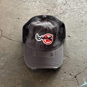 Può includere: Un cappellino trucker grigio antracite con retro in rete nera e visiera usurata con bordi sfilacciati. Il pannello frontale presenta una toppa ricamata con la scritta "björk" in lettere nere su uno sfondo circolare rosso e bianco.
