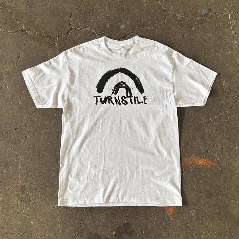 May include: White t-shirt with a black graphic of two arched shapes above the word "TURNSTILE" in a bold, sans-serif font. The shirt is made of a soft material and has a crew neck.