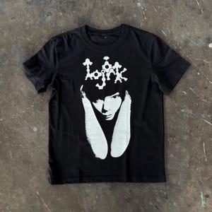 May include: A black short-sleeved t-shirt featuring a white graphic print on the chest. The design displays the text "Lojrk" in a stylised font above a high-contrast, black and white portrait of a person with long, drooping ears or hair.